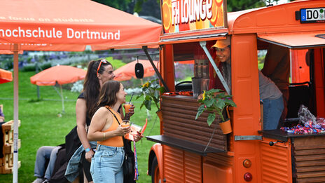 Am Dach eines orangenen Oldtimers ist ein Schild befestigt mit der Aufschrift "FH Lounge". Aus dem Innenraum schaut eine Person nach draußen und lächelt zwei Personen an, die vor einer aufgeklappten Theke des Wagens stehen und unter anderem jeweils einen Becher Slush-Eis halten. Im Hintergrund ist eine Wiese zu sehen mit orangenen Sonnenschirmen.