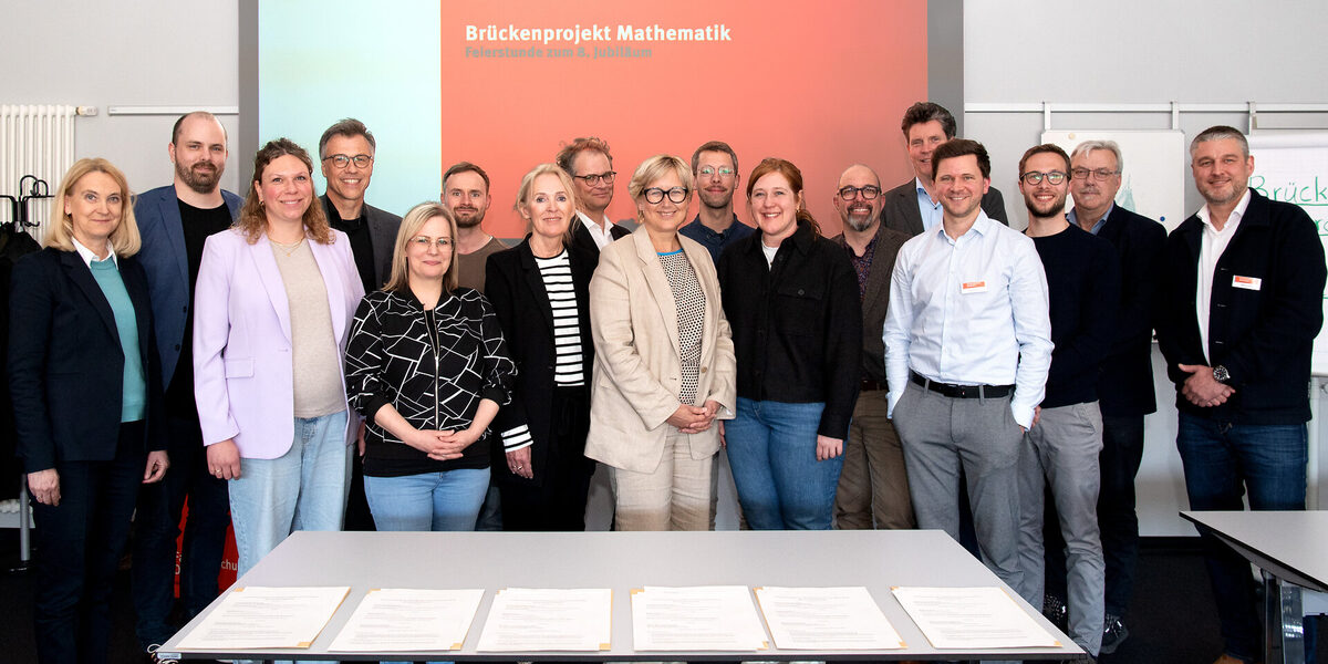 Eine größere Gruppe steht vor einer Beamer-Projektion auf der "Brückenprojekt Mathematik, Feierstunde zum 8. Jubiläum" zu lesen ist.