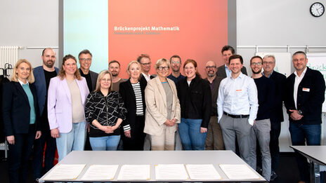 A large group is standing in front of a projector on which the words "Brückenprojekt Mathematik, Feierstunde zum 8. Jubiläum" can be read.
