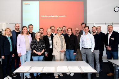 Eine größere Gruppe steht vor einer Beamer-Projektion auf der "Brückenprojekt Mathematik, Feierstunde zum 8. Jubiläum" zu lesen ist.
