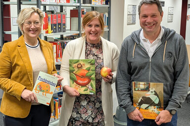 Drei Personen stehen nebeneinander, sie halten jeweils ein Buch vor sich, die Person in der Mitte zusätzlich einen Apfel. Im Hintergrund sind Regale mit weiteren Büchern zu sehen.