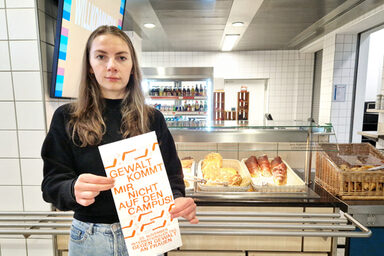 Eine Person steht in der Mensa vor einer Theke mit Backwaren und hält eine Tüte vor sich mit der Aufschrift: "Gewalt kommt mir nicht auf den Campus!".