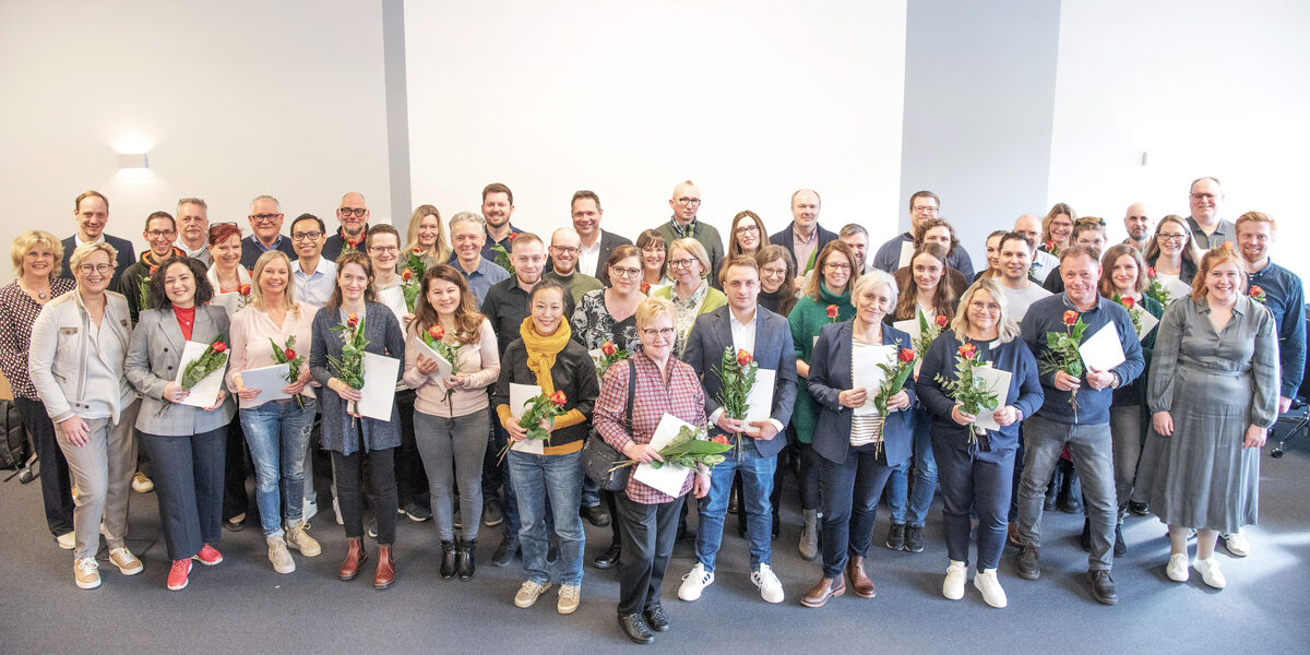 48 people stand in several rows next to each other and look into the camera. Most of them are holding a white envelope and a rose in their hands.