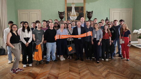 A group of people are standing in a hall, the people in the middle are holding an orange scarf with the words Fachhochschule Dortmund written on it.