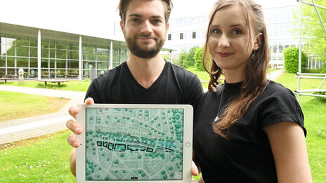 Two people are sitting on the campus of Fachhochschule Dortmund. They are holding a tablet up to the camera. An architectural design can be seen on it.
