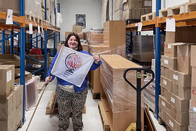 Eine Person hält eine Fahne mit dem Schriftzug "Grenzenlose Wärme" in den Händen. Die Person steht in einem Lager zwischen hohen Regalen, neben ihr ein Hubwagen mit in Folie gewickelten Möbeln.