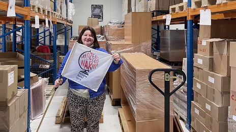 Eine Person hält eine Fahne mit dem Schriftzug "Grenzenlose Wärme" in den Händen. Die Person steht in einem Lager zwischen hohen Regalen, neben ihr ein Hubwagen mit in Folie gewickelten Möbeln.