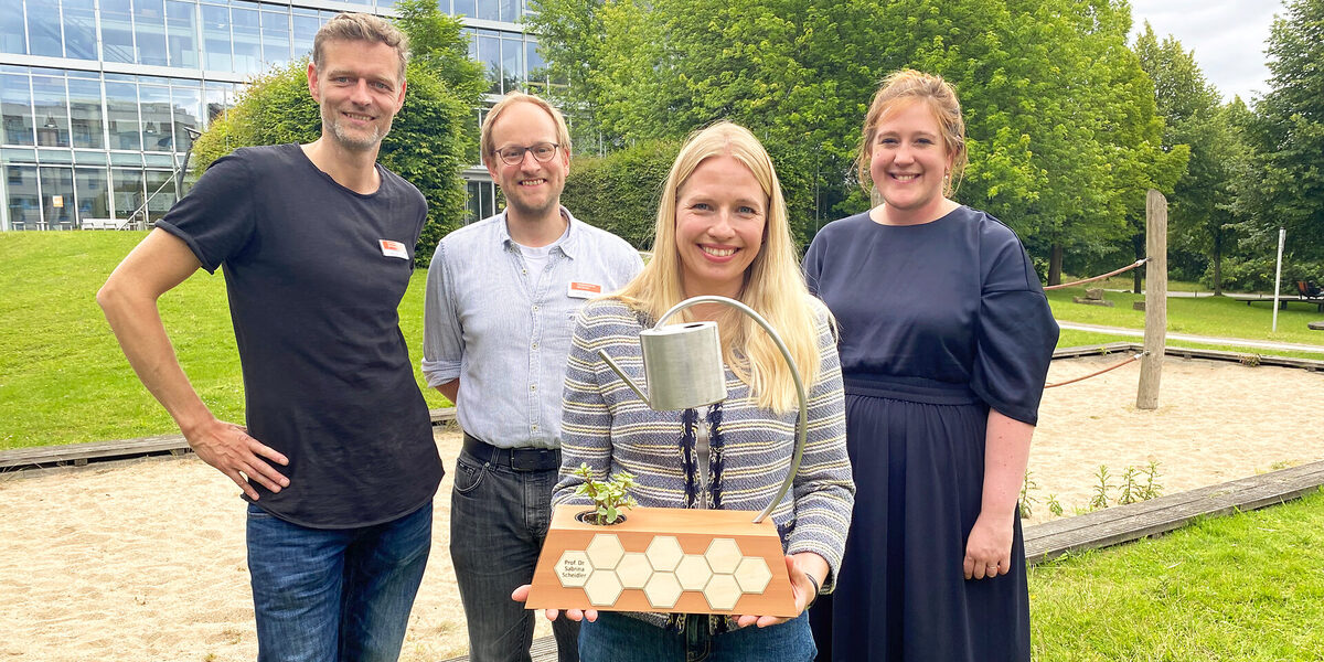 Four people are standing on the Fachhochschule Dortmund campus, one person is holding a prize trophy.