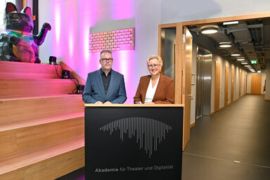 Zwei Personen stehen im Foyer der Akademie für Theater und Digitalität an einem Pult und unterschreiben einen Vertrag.