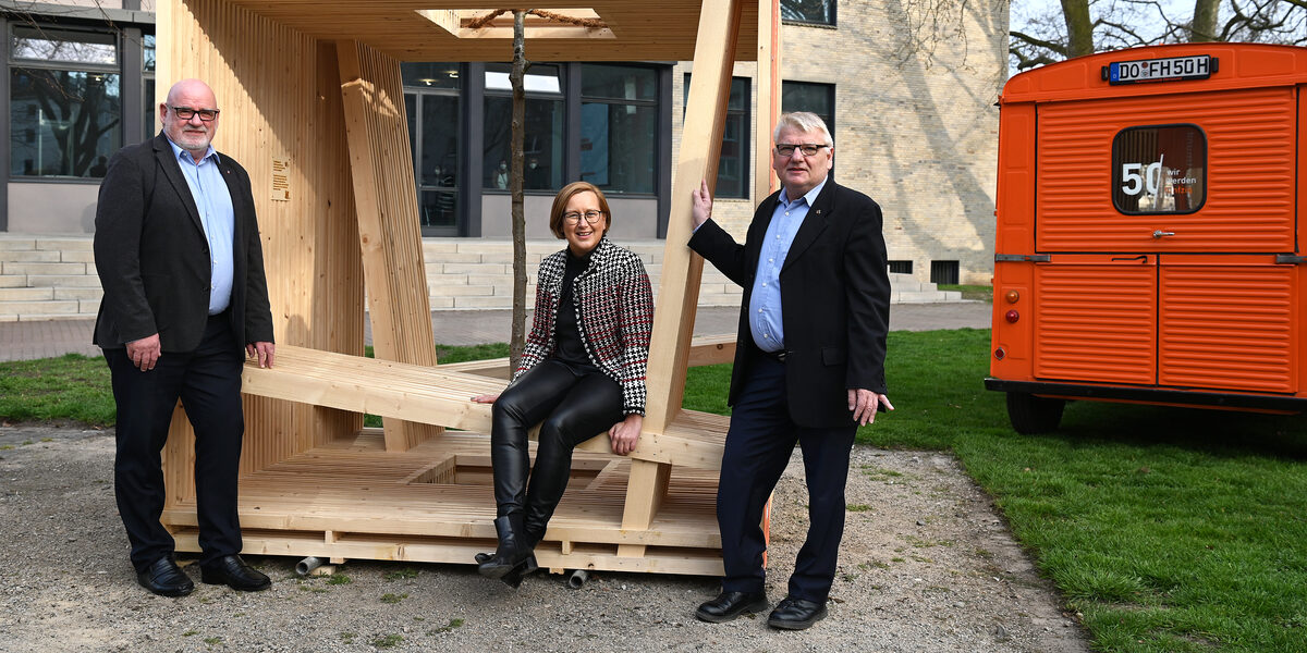 Eine weiblich gelesene Person sitz in einer Würfel-Holzkonstruktion. An den Ecken stehen zwei männlich gelesene Personen. Rechts daneben steht ein orangefarbener Citroen HY.