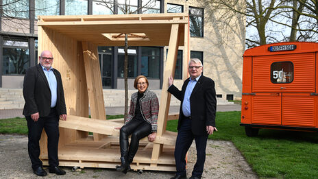 A female read person sits in a wooden cube construction. Two male figures are standing at the corners. To the right is an orange Citroen HY.