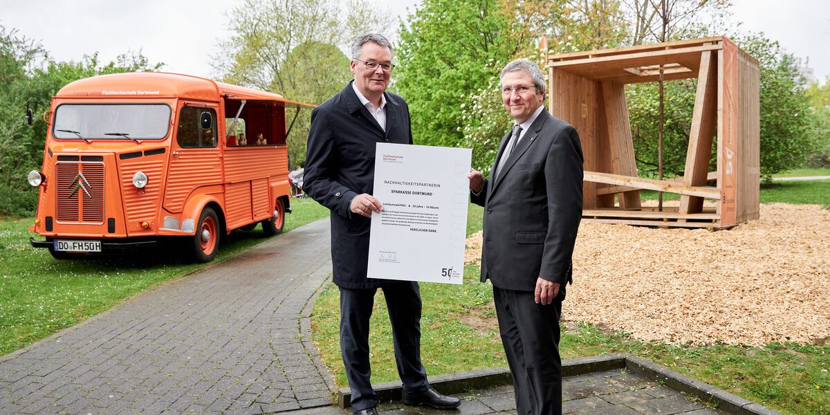 Zwei männlich gelesene Personen stehen vor der Holzkonstruktion eines dreidimensionalen Würfels. Sie halten eine Urkunde in den Händen.