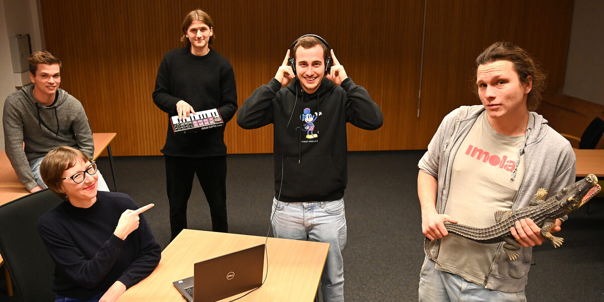 Eine Frau sitzt vor einem Laptop an einem Tisch. Um sie herum stehen vier junge Männer, teilweise mit Musikinstrumenten und Kopfhörern.