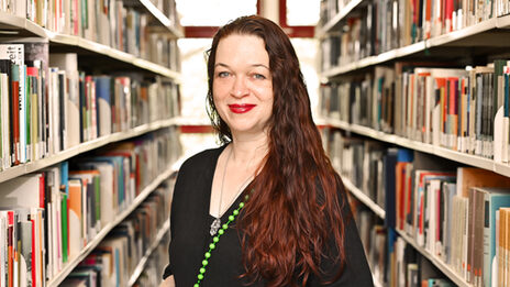 A person in a black top and with long dark red hair stands between bookshelves and looks into the camera.