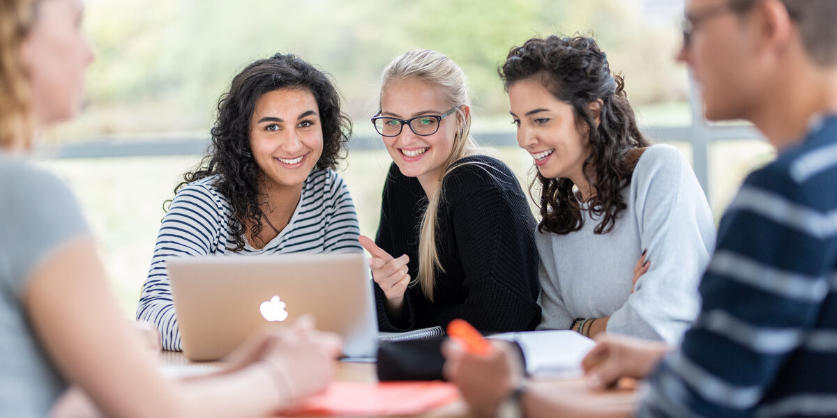 Foto von fünf Studierenden an einem Gruppentisch. Im Fokus drei Studentinnen, die nebeneinander sitzen, vor ihnen auf dem Tisch ein Laptop.