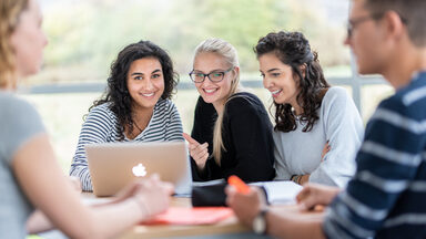 Foto von fünf Studierenden an einem Gruppentisch. Im Fokus drei Studentinnen, die nebeneinander sitzen, vor ihnen auf dem Tisch ein Laptop.