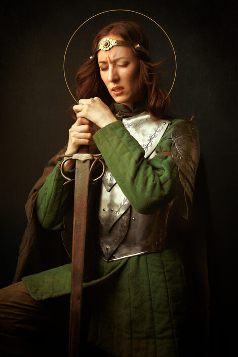 Eine Person mit langem rotem Haar vor schwarzem Hintegrund in einer Ritterrüstung stützt sich auf ein Schwert, hält die Augen geschlossen und hat die Stirn in Falten gelegt.