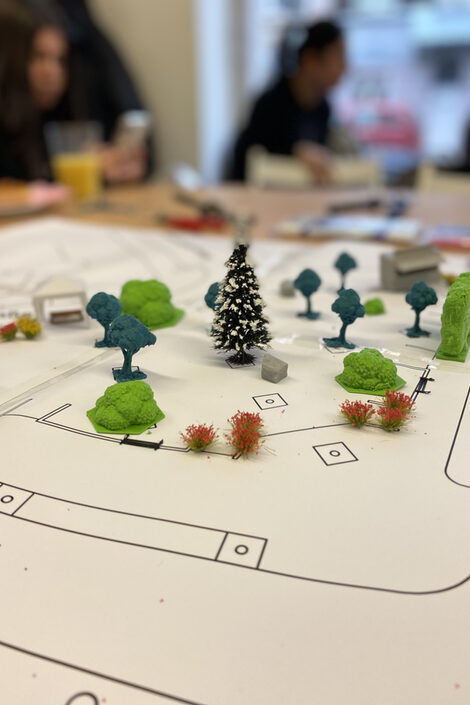 The foreground shows a simple site plan with model trees standing on it. The blurred background shows people sitting at a table.