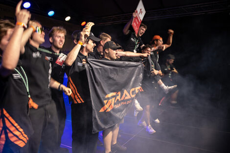 Several people cheer joyfully with their arms raised, two of them holding a tarpaulin with the Race-Ing. team's logo on it.