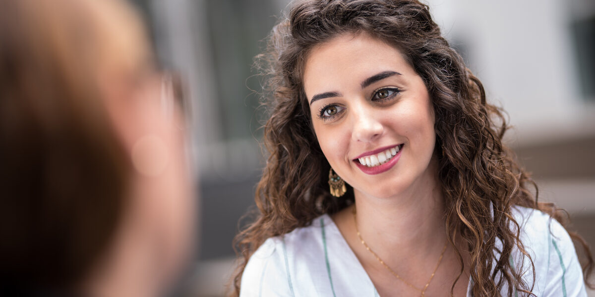 Closeup of a young woman with dark curly hair smiling at a woman sitting across from her.__Closeup of a young woman with dark curly hair smiling at a woman sitting across from her.
