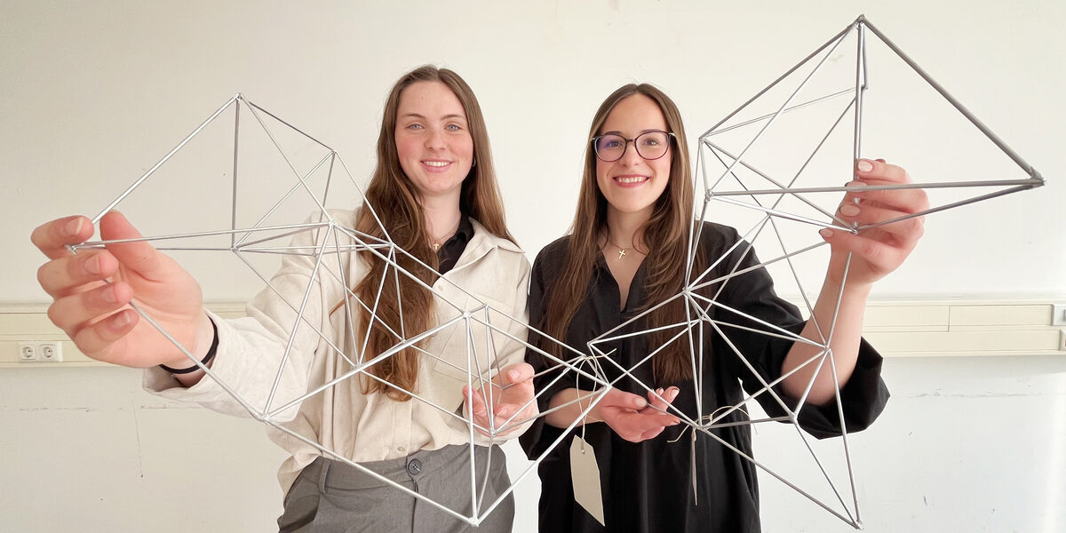 Two smiling people each hold a delicate structure made of metal rods up to the camera.