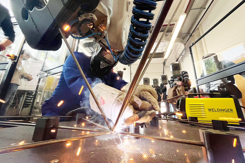 In the foreground, the ends of several metal rods touch. A person crouches behind them and welds them together. A part of a robot arm holding one of the rods protrudes into the picture from above. In the background, several people wearing protective masks stand and watch.