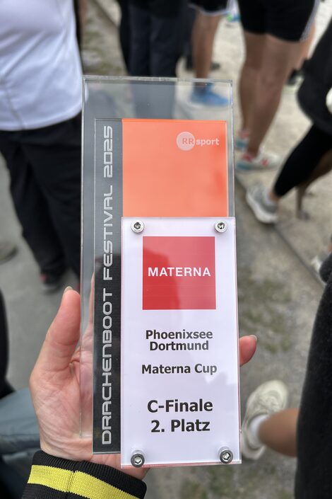 Eine Hand hält einen Pokal aus Plexiglas und Kunststoff mit der Aufschrift "Materna. Phoenixsee Dortmund. Materna Cup. C-Finale, 2. Platz". Drum herum sind die Beine mehrerer Menschen zu sehen.