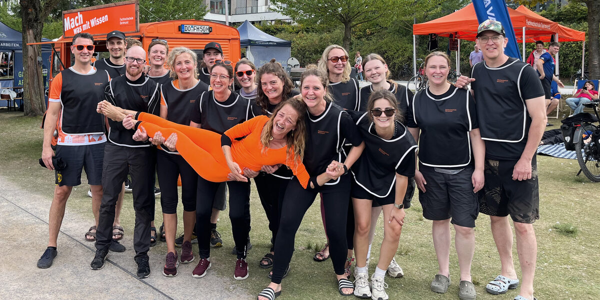15 Personen in schwarzen Trikots tragen eine weitere Person in Orange auf Händen, stehen nebeneinander und schauen in die Kamera. Im Hintergrund stehen ein Auto und zwei Zelte, beide orange, auf einer Wiese.
