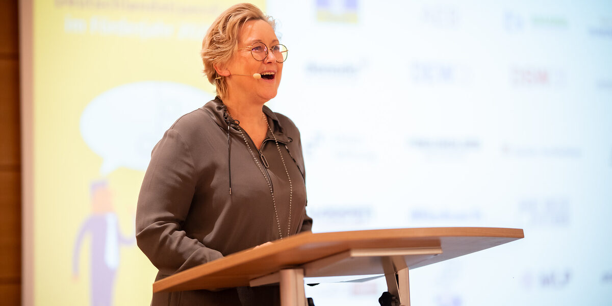 Eine Person mit Kopfmikrofon steht an einem Rednerpult und spricht. Im Hintergrund ist eine Projektion zu sehen.