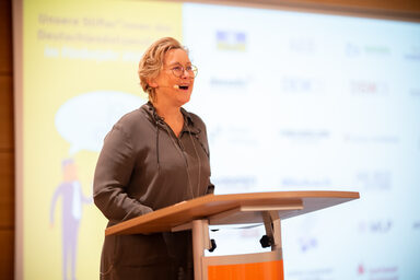 Eine Person mit Kopfmikrofon steht an einem Rednerpult und spricht. Im Hintergrund ist eine Projektion zu sehen.