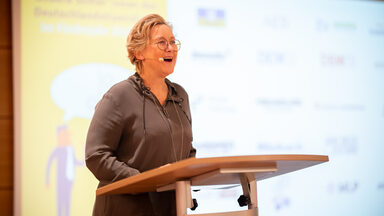 Eine Person mit Kopfmikrofon steht an einem Rednerpult und spricht. Im Hintergrund ist eine Projektion zu sehen.