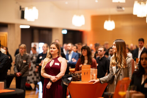 Viele festlich gekleidete Personen stehen, nach vorn links gewandt, in einem festlichen Raum und hören vermutlich einer Person zu, die zu ihnen spricht.