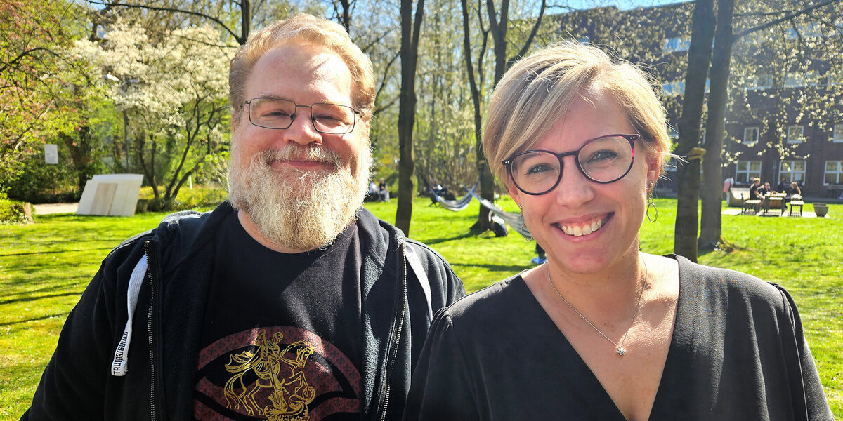 Zwei Personen stehen im Sonnenschein in einem grünen Innenhof und blicken freundlich in die Kamera.