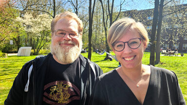 Zwei Personen stehen im Sonnenschein in einem grünen Innenhof und blicken freundlich in die Kamera.