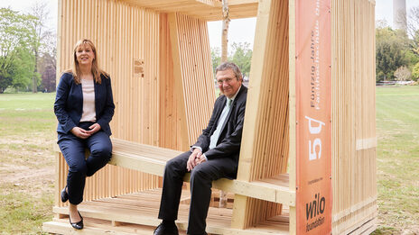 Two people are sitting in a large wooden cube. The Westfalenpark television tower can be seen in the background.