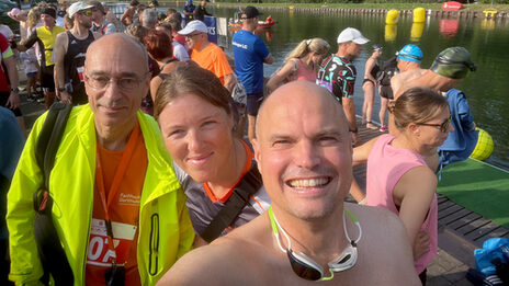 Selfie von drei Personen unter freiem Sommerhimmel mit vielen weiteren Personen im Hintergrund am Ufer eines Wasserlaufs.