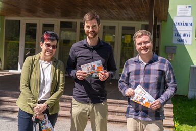 Three people are standing in front of a school holding flyers from Fachhochschule Dortmund.