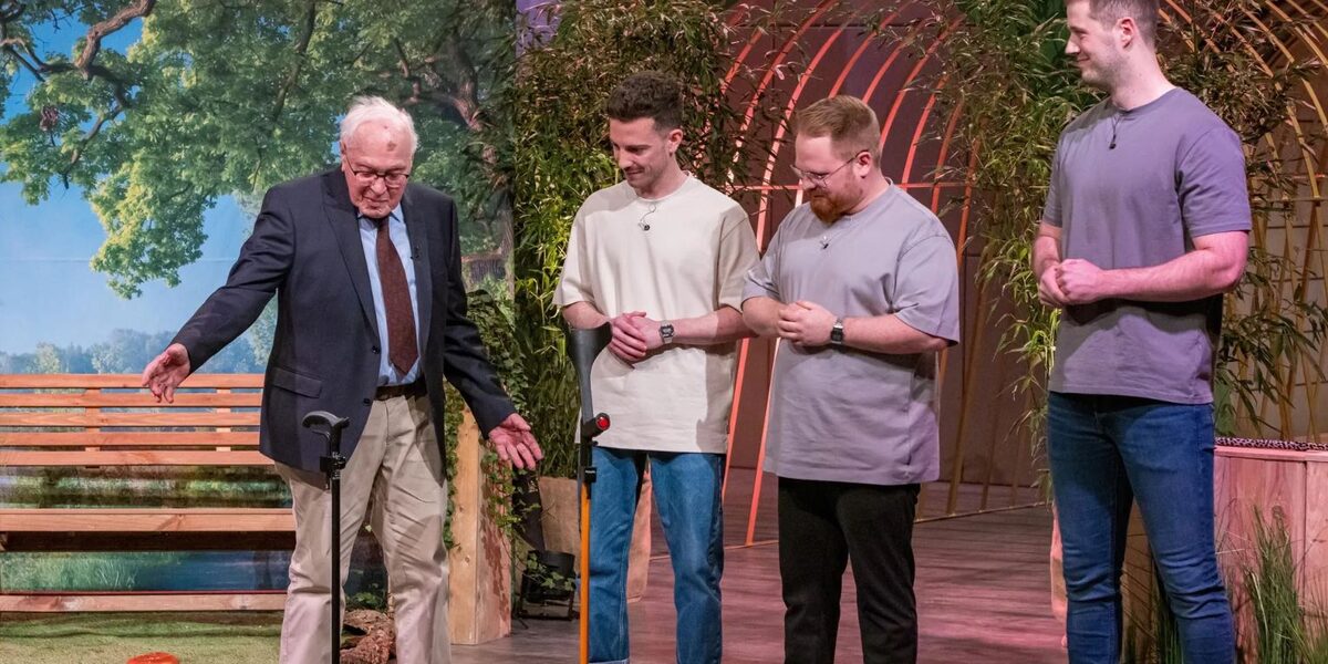 Ein älterer Mann zeigt, wie sein Gehstock allein stehen bleibt. Drei junge Männer schauen dabei zu.