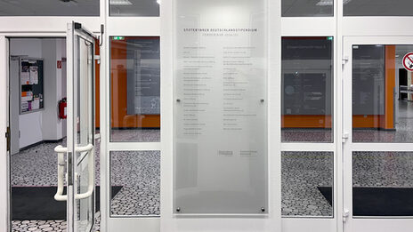 A gray, semi-transparent, roughly door-sized plaque with the names of companies hangs on a pillar between two entrance doors to a foyer room.