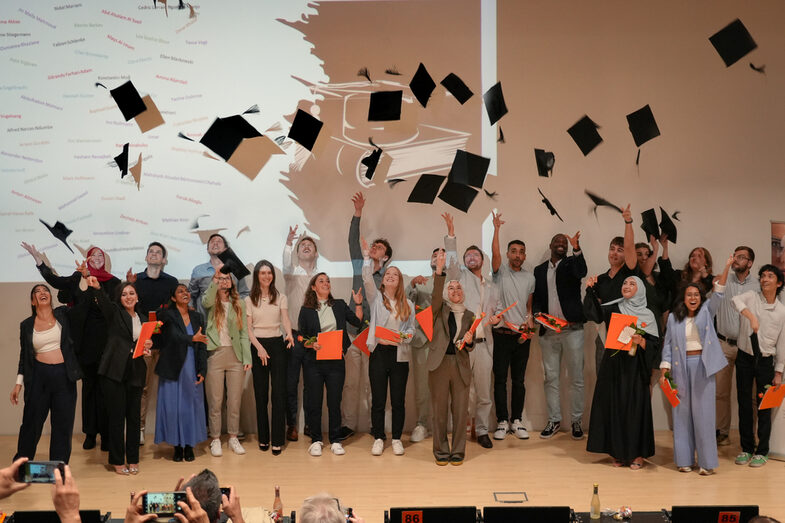 A group of people stand on a stage and throw black doctor's hats in the air to symbolize graduation. The people wear different clothes, including suits, dresses and casual outfits. Some are holding orange document folders. A white wall with an abstract pattern and texts can be seen in the background. Spectators with cameras can be seen in front of the stage taking photos.