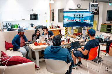 Five people sit in a room at a table with laptops and technical equipment, next to a large screen showing a virtual room with circle symbols.