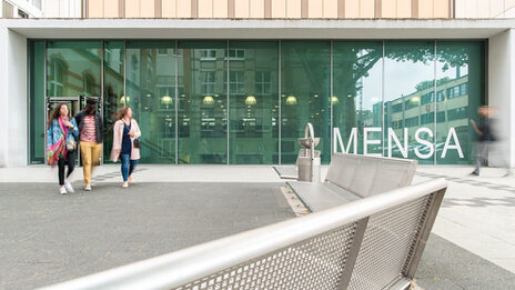 Three students come out of the new canteen on Sonnenstrasse. The glass façade of the canteen is large in the picture.