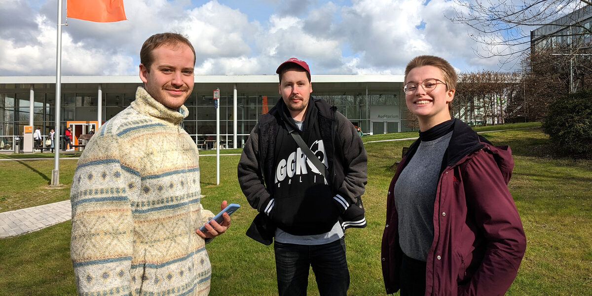 Drei Personen stehen auf einer Wiese auf dem FH-Campus an der Emil-Figge-Straße und sprechen miteinander.