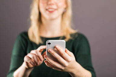 Young woman - head at the top of the crop - holding a smartphone in her left hand in front of her, almost touching the display with the index finger of her right hand.