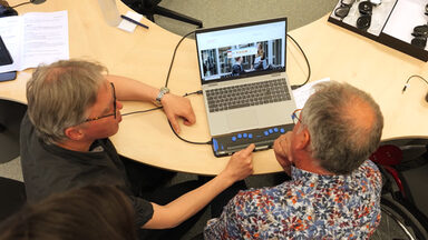 Zwei Personen sitzen an einem Tisch. Und schauen auf einen Laptop. Eine Person zeigt der anderen eine Vorrichtung, mit denen blinde Menschen die Inhalte des Bildschirms erfassen können.