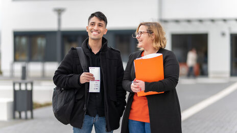 Photo of an employee and a student standing in front of a campus building. She is holding an orange folder under her arm, he has a flyer in his hand. They are both laughing.
