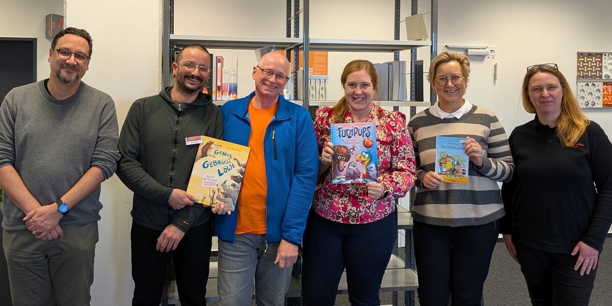 Six people with a few children's books in their hands stand in a room and look into the camera.