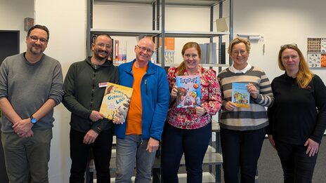 Sechs Personen mit einigen Kinderbüchern in den Händen stehen in einem Raum und schauen in die Kamera.