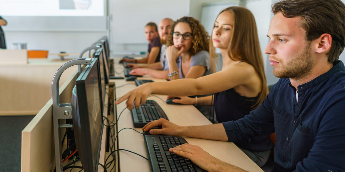 Foto von fünf Studierenden in einer Reihe an PCs. Eine Studentin zeigt den anderen etwas auf dem Bildschirm ihres Sitznachbars.__Reihe an fünf PCs und schauen auf die Bildschirme.__Five students sit in a row at five PCs. One student is showing something to the others on the screen of the student sitting next to her.