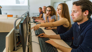 Foto von fünf Studierenden in einer Reihe an PCs. Eine Studentin zeigt den anderen etwas auf dem Bildschirm ihres Sitznachbars.__Reihe an fünf PCs und schauen auf die Bildschirme.__Five students sit in a row at five PCs. One student is showing something to the others on the screen of the student sitting next to her.